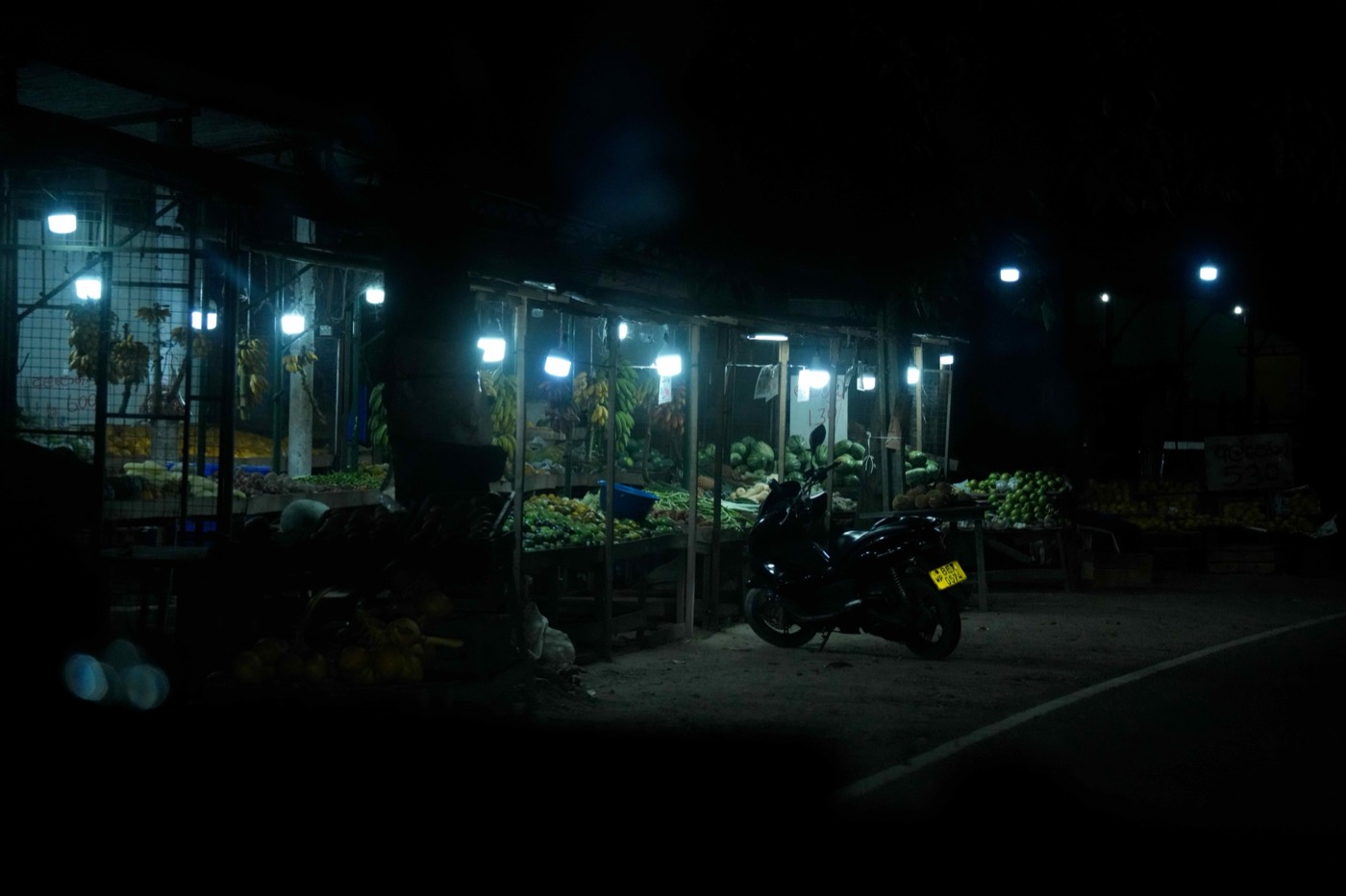 闇夜に浮かび上がる路上の青果マーケット。バナナや野菜がずらりと並ぶ