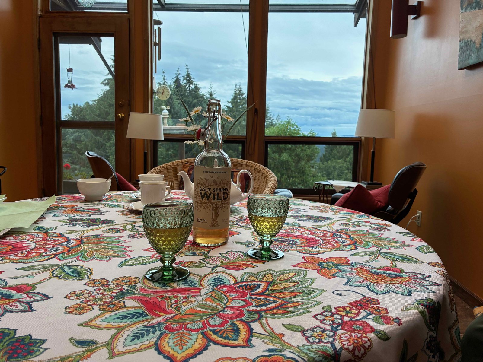 窓辺のテーブルに置かれたSalt Spring Wild Ciderのボトルとグラス。窓の外にハチドリの給餌器が見える